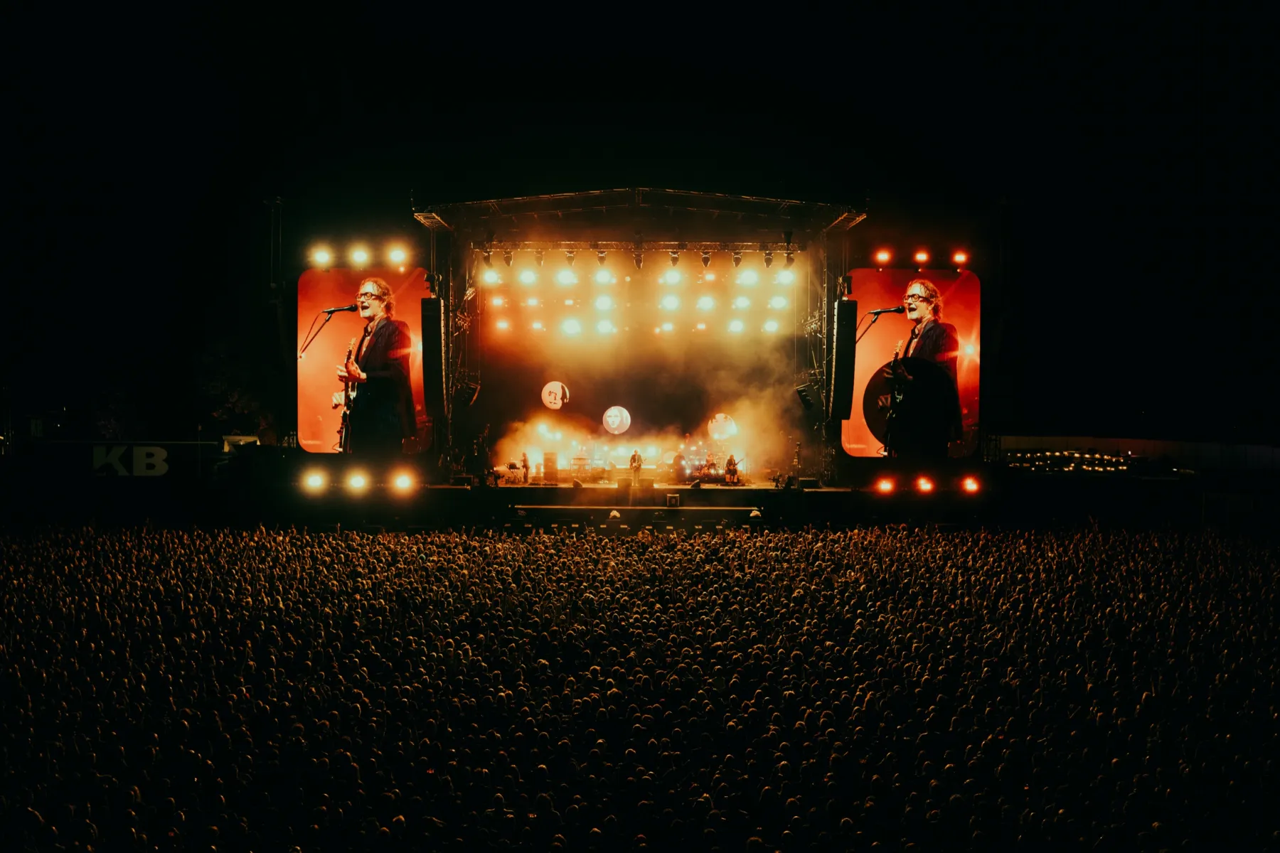 A festival crowd at night in front of a main stage lit up in orange