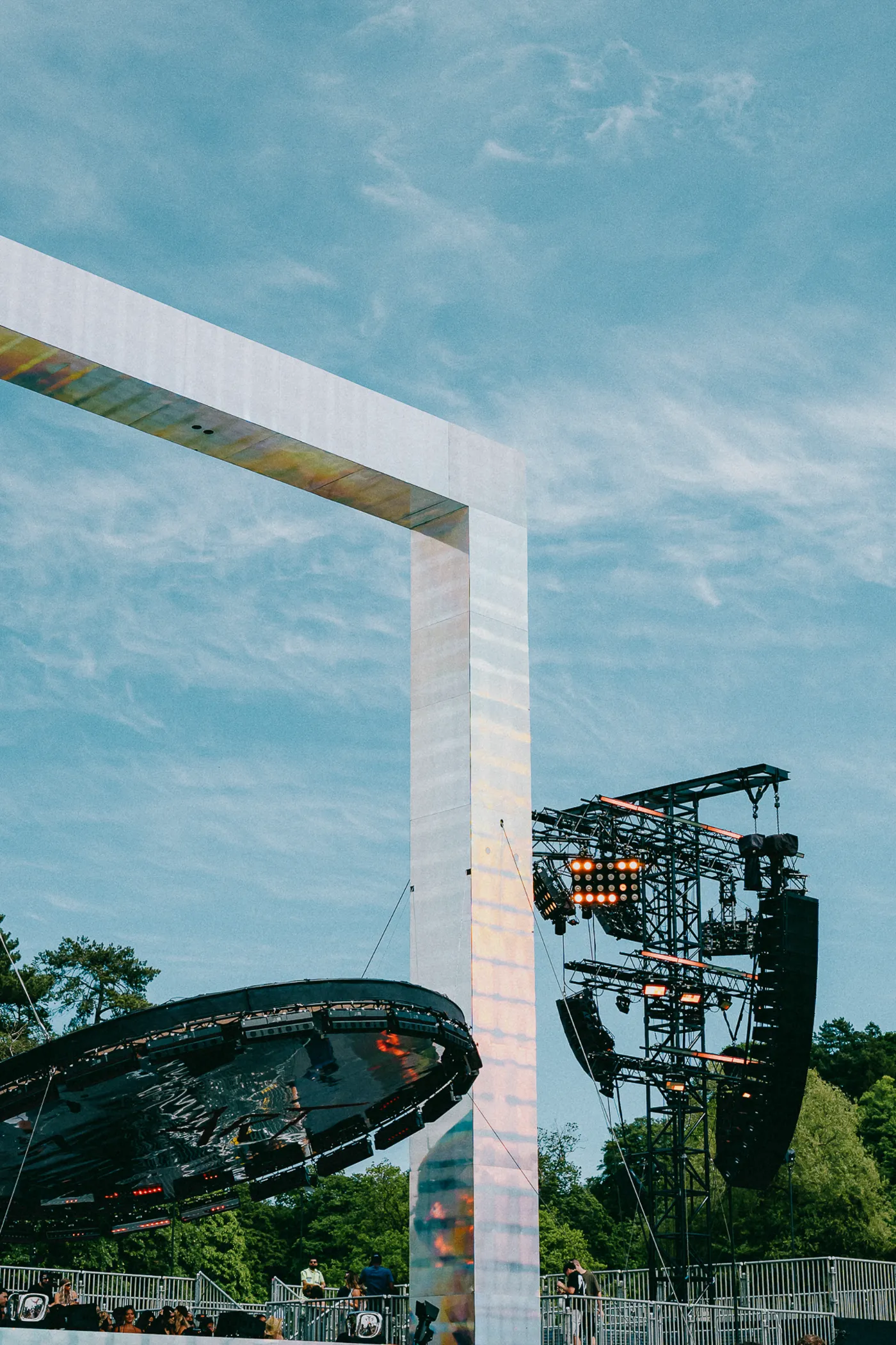 A tall mirrored arch, lighting towers and a round stage roof under a blue sky on a festival site